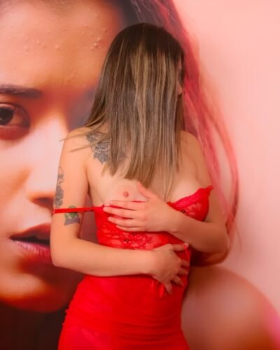 Scort en Temuco Luisa con un babydoll rojo posando enigmática frente una una pared con un rostro de mujer lista para tener sexo contigo en Temuco